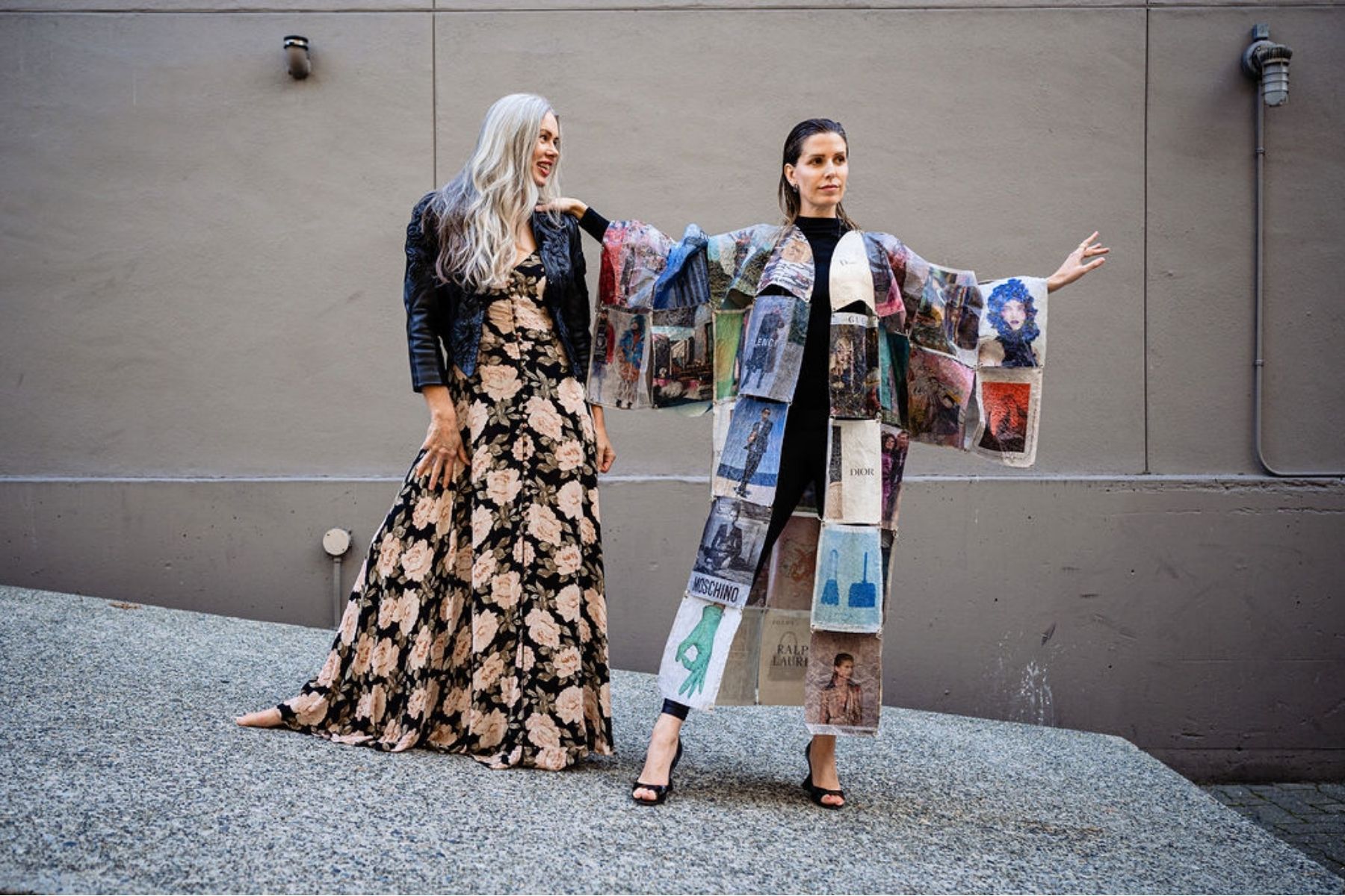 Two women posing outdoors against a grey wall. The woman on the right wears a wide-sleeved kimono made using a Momigami technique from fashion ads, standing tall in a black outfit. The woman on the left wears a floral maxi dress and a black leather jacket.