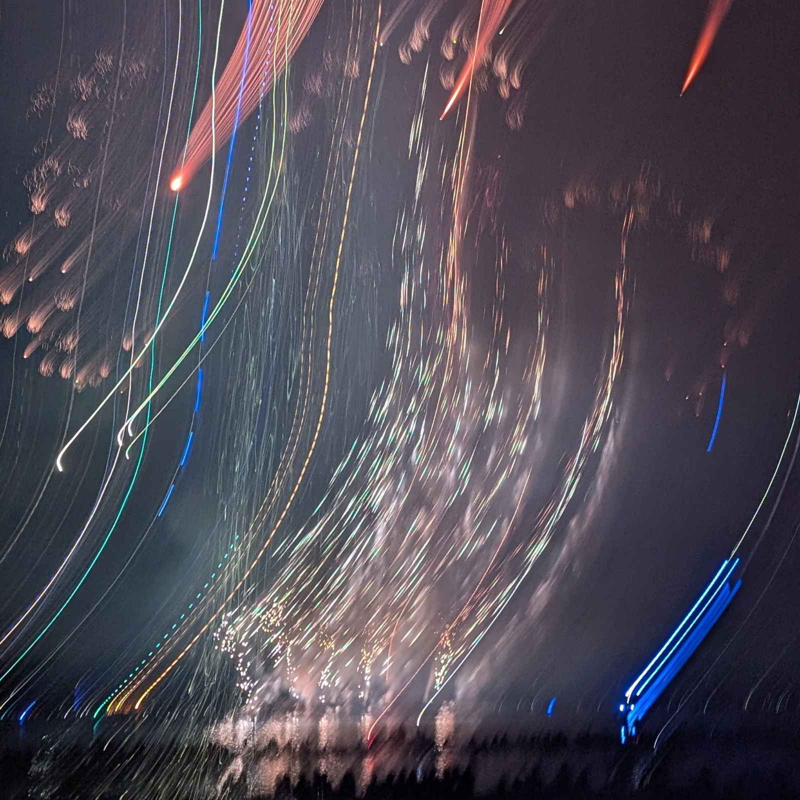 An abstract aesthetic photograph capturing the long, colorful light trails of multiple fireworks exploding against a dark sky.